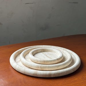 Puede incluir: Tres bandejas redondas de mármol blanco, anidadas, de diferentes tamaños. Las bandejas están hechas de piedra natural y tienen una textura ligeramente rugosa.