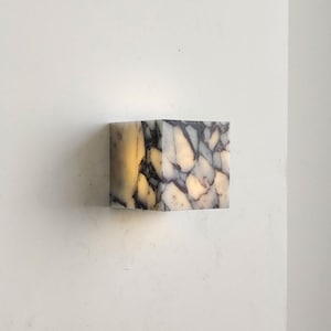 May include: A square marble wall light with a distinctive pattern of white, grey, and black veining. The light is illuminated internally, emitting a warm glow. The fixture is mounted on a white wall.