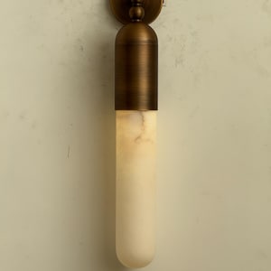 May include: A wall-mounted light fixture with a cylindrical design. The fixture features a bronze-colored metal top and a long, off-white, marble-like bottom. The light is designed to be mounted on a wall.
