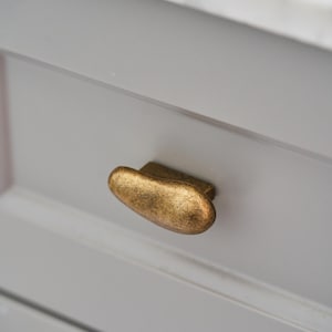 May include: A brass-toned cabinet knob with an organic, pebble-like shape and a textured, weathered finish. The knob is mounted on a light gray drawer front, which features a horizontal recessed panel design and a smooth, matte surface texture.