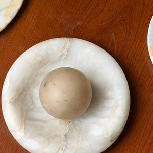 May include: A round, white marble dish with a beige sphere in the center. The dish has a circular shape with a slightly raised edge and a natural veining pattern. The sphere is smooth and solid.