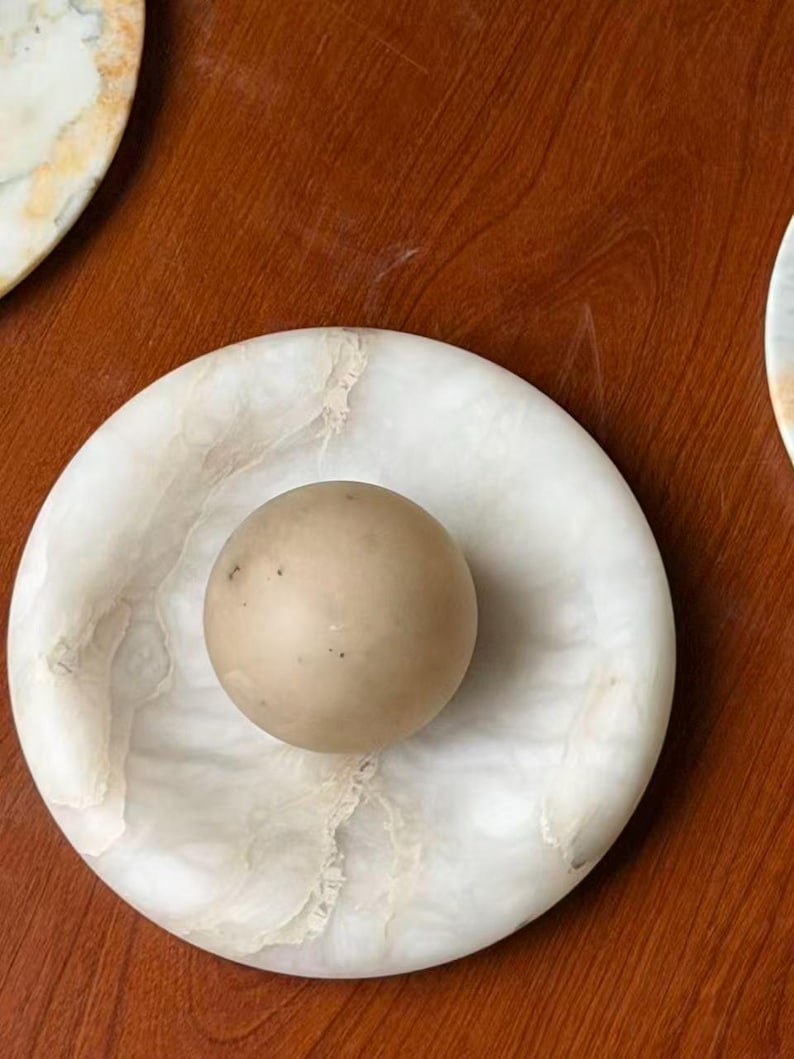 May include: A round, white marble dish with a beige sphere in the center. The dish has a circular shape with a slightly raised edge and a natural veining pattern. The sphere is smooth and solid.