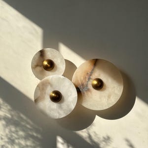 May include: Three circular wall art pieces made of white and tan marble with brass center accents. The pieces are arranged in a cluster on a white wall, with shadows cast across the surface.
