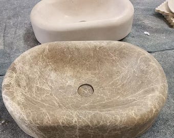 May include: Two oval stone sinks. The front sink is a mottled brown with a central drain hole. Behind it is a lighter-coloured sink, also oval, with a drain hole. A small, rounded stone object is behind the lighter sink.