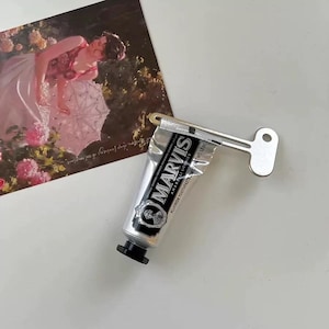 Pu&ograve; includere: Un tubetto di dentifricio Marvis Amarelli Licorice con uno spremi-tubo a forma di chiave in metallo argentato. Il tubetto di dentifricio &egrave; argentato con testo nero e un tappo nero. Sullo sfondo una cartolina con un dipinto di una donna in abito rosa.