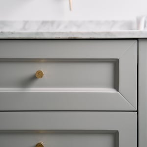 Peut inclure: Gros plan sur une vanité de salle de bain moderne avec deux tiroirs gris et des éléments dorés. Les tiroirs ont des boutons dorés texturés et sont posés contre un comptoir en marbre blanc et des accessoires muraux.