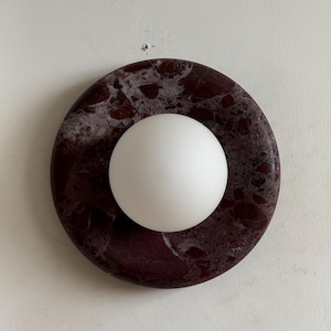 May include: A round, dark red marble wall sconce with a white orb light fixture. The marble has a natural, veined pattern. The light is a smooth, opaque white sphere.