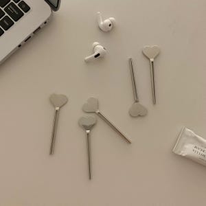 Pu&ograve; includere: Una collezione di cucchiai argentati a forma di cuore e un paio di auricolari wireless bianchi sono disposti su una superficie di colore chiaro. &Egrave; visibile anche un tubetto di Crema Camelia. Una tastiera di un laptop &egrave; parzialmente visibile.