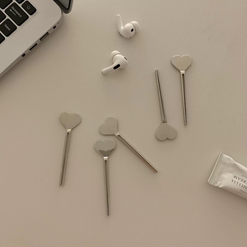 Pu&ograve; includere: Una collezione di cucchiai argentati a forma di cuore e un paio di auricolari wireless bianchi sono disposti su una superficie di colore chiaro. &Egrave; visibile anche un tubetto di Crema Camelia. Una tastiera di un laptop &egrave; parzialmente visibile.