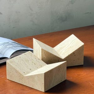 May include: Two beige stone bookends with a geometric design. Each bookend has a rectangular base and a slanted top, creating a V-shape. The bookends are made of a porous stone material and sit on a wooden surface.