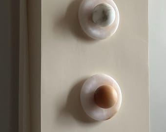 May include: Two decorative wall sconces. Each sconce features a circular, off-white base with a central, spherical accent. One accent is white and gray, the other is a warm brown. The sconces are mounted on a light beige wall.