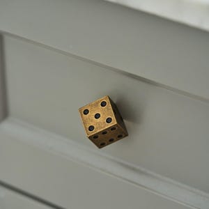 Op de afbeelding: Een kubusvormige meubelknop van antiek messing met een donkere, verweerde afwerking. De knop is ontworpen als een zeszijdige dobbelsteen, met verzonken zwarte cirkelvormige stippen op de zichtbare zijden, gemonteerd op een lichtgrijze ladefront.