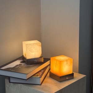 Puede incluir: Dos lámparas cúbicas iluminadas con un brillo cálido, apoyadas sobre bases oscuras. Una lámpara está sobre una pila de libros, la otra sobre un bloque de hormigón. Las lámparas están hechas de un material translúcido, creando una luz ambiental suave.