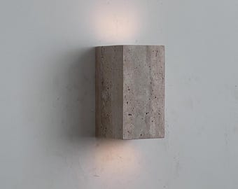 May include: A rectangular, light gray wall sconce with a textured surface. The sconce has a geometric design and emits a soft, warm light from the top and bottom. The fixture is mounted on a white wall.
