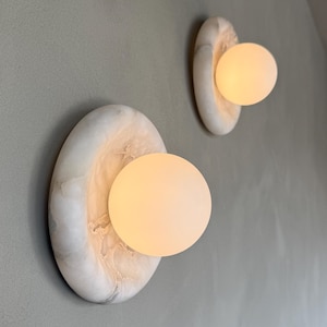 Könnte beinhalten: Zwei Wandleuchten mit einem einzigartigen Design. Jede Leuchte hat eine ovale Basis aus weißem Marmor mit einem warmen, kugelförmigen Licht. Die Leuchten sind an einer hellgrauen Wand montiert und schaffen eine moderne Ästhetik.