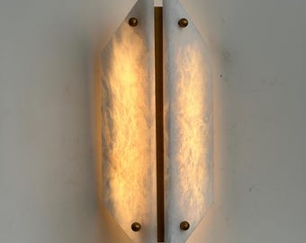 May include: A modern wall sconce with a geometric design. The sconce features two elongated, angled panels of white, translucent material, possibly alabaster or a similar stone, with a warm, glowing light emanating from within. The panels are mounted on a brass-colored backing and secured with small, round brass accents.