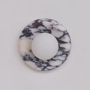 May include: A circular wall light featuring a white marble base with dark purple and black veining. A smooth, matte white spherical glass globe is centred on the marble disc, creating a contrast between the textured stone and the plain orb.