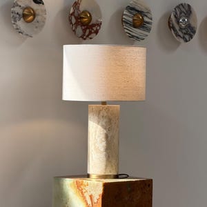 May include: A beige stone lamp with a rectangular base and cylindrical body, topped with a rectangular lampshade. Decorative marble wall accents with brass hardware are visible in the background.