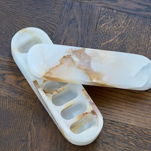 May include: An elongated, oval-shaped white and tan stone tray with four recessed compartments and a matching removable lid. The natural stone features translucent white areas with swirling brown and gold veining throughout the polished surface.