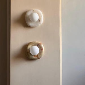 May include: Two wall sconces with a natural stone appearance. The top sconce has a white and cream marbled design, while the bottom one features a beige, brown, and red pattern. Each sconce has a round, white light bulb in the centre.