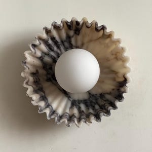 May include: A marble bowl with a ruffled edge, containing a white sphere. The bowl's colour scheme is cream and grey, with dark grey veining. The sphere is smooth and white, contrasting with the textured bowl. The bowl's diameter is approximately 15 cm.