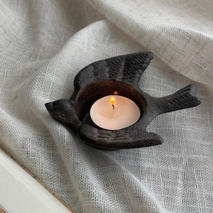 May include: A black cast iron bird-shaped candle holder with a lit white tea light candle. The candle holder has detailed wing and tail features. The flame glows brightly against the dark metal.
