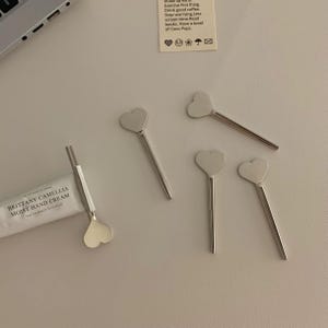 Pu&ograve; includere: Un assortimento di agitatori argentati a forma di cuore e un tubetto di crema per le mani Brittany Camellia Moist sono disposti su una superficie chiara. Gli agitatori hanno un manico lungo e sottile. Il tubetto di crema per le mani &egrave; bianco con testo nero.