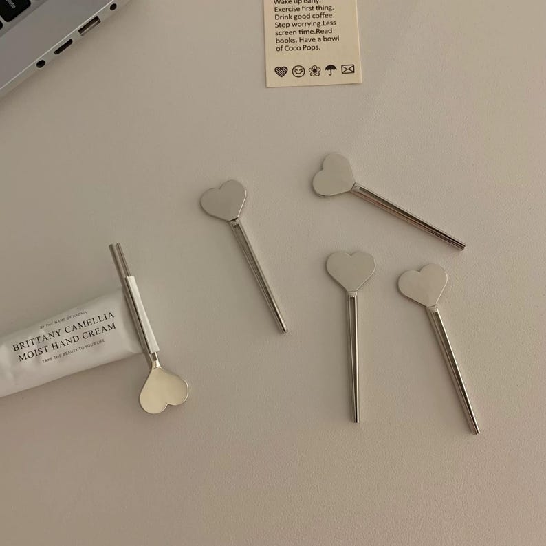 Pu&ograve; includere: Un assortimento di agitatori argentati a forma di cuore e un tubetto di crema per le mani Brittany Camellia Moist sono disposti su una superficie chiara. Gli agitatori hanno un manico lungo e sottile. Il tubetto di crema per le mani &egrave; bianco con testo nero.