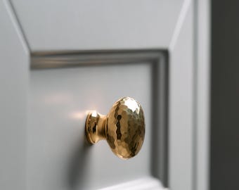 May include: A close-up of a gold-toned cabinet knob with a hammered finish. The knob is mounted on a white cabinet door.
