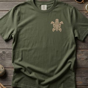 May include: An olive green short-sleeve t-shirt featuring a light tan graphic of a stylized turtle on the left chest area. The shirt has a crew neckline and a visible interior neck label with the text Comfort Colors and size L.