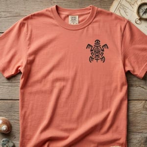 May include: A coral-colored short-sleeve t-shirt featuring a black graphic of a stylized turtle on the upper left chest. The inner neck label displays the text Comfort Colors and the size L.