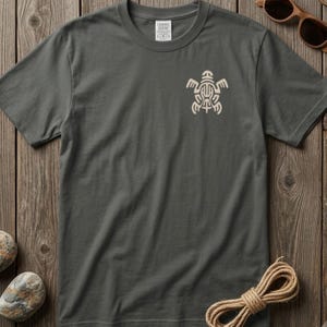 May include: A dark gray short-sleeve t-shirt featuring a cream-colored geometric turtle graphic on the left chest. The shirt has a crew neckline and a visible interior neck label with the text Comfort Colors, indicating a size L/G/G.