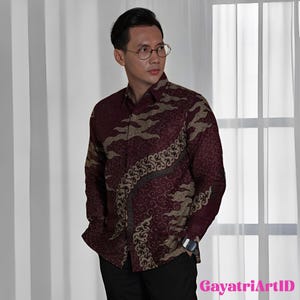 Peut inclure: Chemise bordeaux à motifs batik beiges et marron foncé. La chemise à manches longues a un col classique. Le texte "GayatriArtID" est visible en bas à droite.
