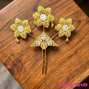 May include: A gold-colored hair accessory set featuring three openwork flower designs with clear crystal centers and dangling leaf accents. The set includes a central triangular piece with crystals and a two-pronged hairpin.