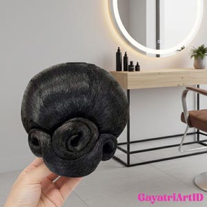 May include: A black hair bun with a rolled, decorative design. The hairpiece is held in a hand, with a salon-style vanity and mirror in the background. The text "GayatriArtID" is visible in the bottom right corner.