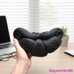 Könnte beinhalten: Ein schwarzes Haarnetz, das in einer Hand gehalten wird. Das Haarnetz besteht aus einem feinen, gewebten Material und dient dazu, das Haar an Ort und Stelle zu halten. Der Hintergrund umfasst einen Laptop, eine Brille und eine kleine Topfpflanze auf einem Holztisch.