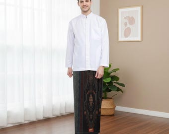 May include: A man wearing a white long-sleeved shirt and a black patterned sarong. He is also wearing a black cap. The sarong has a dark brown and black pattern. The man is standing in front of a window.