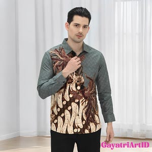May include: A long-sleeved button-down shirt with a batik design. The shirt features a gray-green color on the sleeves and back, with a cream and brown batik pattern on the front. The shirt is paired with black pants.