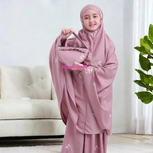 Może przedstawiać: Osoba trzyma różowy strój do modlitwy, składający się z nakrycia głowy, tuniki i spódnicy. Strój zdobią białe, haftowane wzory kwiatowe. Widoczna jest również pasująca torba. Strój wykonany jest z miękkiej, zwiewnej tkaniny.