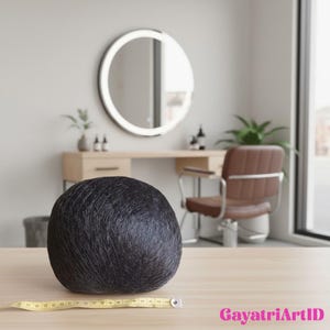 May include: A large, dark grey ball of yarn rests on a light wood surface, with a yellow measuring tape in front. The background features a circular, illuminated mirror, a vanity, and a brown leather chair, suggesting a hair salon or craft studio setting.