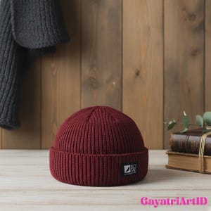 May include: A burgundy ribbed knit beanie with a small rectangular label on the front. The hat is displayed on a light wood surface with a wooden background. A gray scarf hangs in the background.
