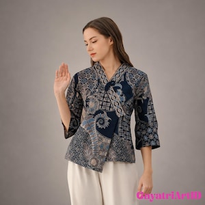 Puede incluir: Una blusa cruzada azul marino con un estampado floral batik en tonos azules, grises y blancos. La blusa tiene mangas tres cuartos y un diseño asimétrico. La modelo lleva pantalones blancos.