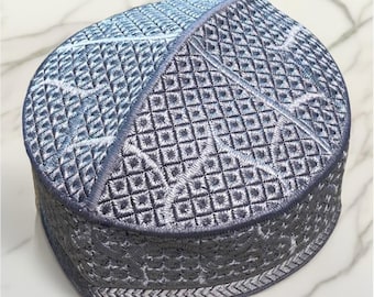 May include: A gray and blue embroidered skullcap with a geometric pattern. The cap features a diamond-like design with intricate stitching and a decorative border. The cap is a traditional head covering.