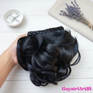 May include: A black hairpiece with wavy curls and a braided top section. The hairpiece is held in a hand, with a small bowl of hairpins and a bundle of lavender in the background.
