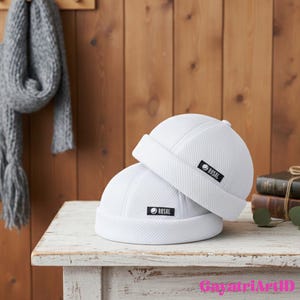 Op de afbeelding: Twee witte skull caps gestapeld op een verweerde witte tafel. Elke cap heeft een klein zwart label met het woord "ROSAL" in wit. Een grijze sjaal hangt op de achtergrond aan een houten muur.