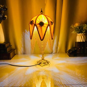 May include: A decorative table lamp with a yellow lampshade and a gold-coloured base. The lampshade has a scalloped edge with beaded fringe. The base has an ornate floral design. The text "Bright freedom" is visible on the surface.