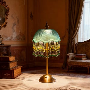 Pode incluir: Uma luminária de mesa de estilo vintage com uma cúpula de vidro verde-azulado e franjas de contas. A luminária tem uma base e haste de latão, e a cúpula é adornada com contas penduradas em tons de amarelo e verde. O texto "Bright freedom" está na mesa.