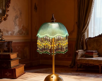 Puede incluir: Una lámpara de mesa de estilo vintage con una pantalla de cristal verde azulado y flecos de cuentas. La lámpara tiene una base y un tallo de latón, y la pantalla está adornada con cuentas colgantes en tonos amarillos y verdes. El texto "Bright freedom" está en la mesa.