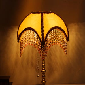 May include: A vintage-style table lamp with a golden yellow lampshade. The shade is adorned with a dark brown trim and hanging beaded fringe. The lamp has a decorative, ornate gold-colored base and a pull chain.