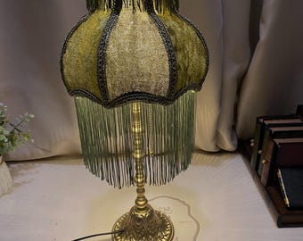 May include: A vintage-style table lamp with a green fringed lampshade. The shade has a scalloped design with dark trim. The lamp base is gold-colored with an ornate, floral design. The text "Bright freedom" is visible on the surface.